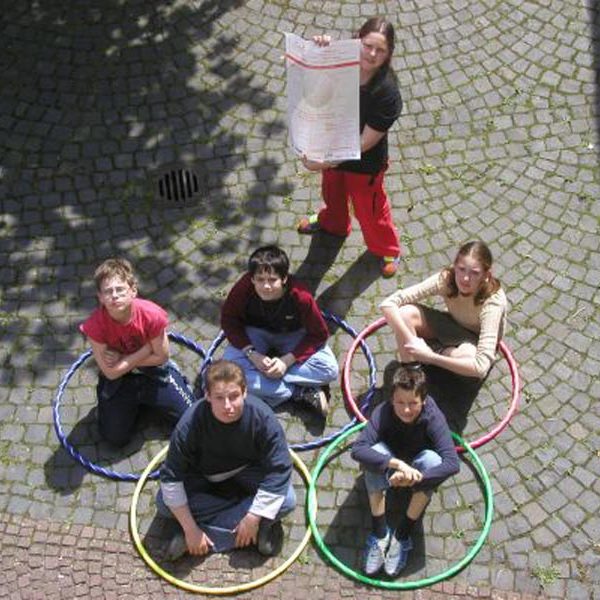 Projekt "Netzolympiade 2004". Fünf Kinder sitzen auf dem Boden in Hula Hoop Reifen, die wie die olympischen Ringe angeordnet sind. Daneben steht ein weiteres und hält ein Plakat in die Kamera.