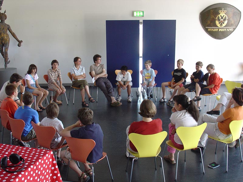 Projekt "Klickerkids - Internetzeitung von und für Kinder". Kinder und Erwachsene sitzen in einem Stuhlkreis.