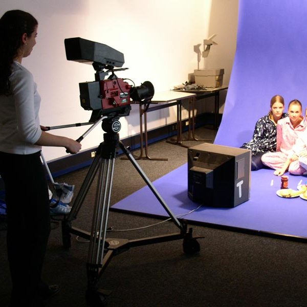 Projekt "Geld, Macht, glücklich - Fächerübergreifendes Medienprojekt". Drei Mädchen sitzen auf dem Boden vor einem Blue-Screen. Vor ihnen steht eine Frau mit Filmkamera.