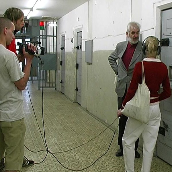 Projekt "DDR Zeitzeugen". Drei junge Erwachsene und ein älterer Herr stehen vor einer Gefängniszellentür. Sie sind mit Filmkamera und Kopfhörern ausgestattet und filmen den älteren Herren.