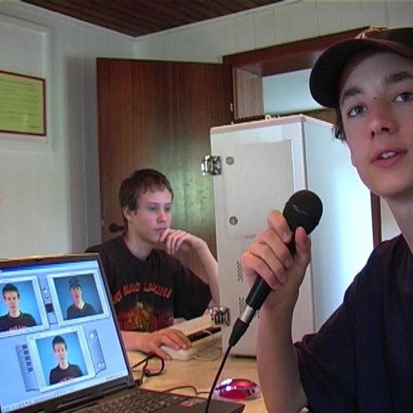 Projekt "Don´t believe what you see". Ein Junge sitzt vor einem Laptop und hält ein Mikrofon in der Hand. Auf dem Bildschirm des Laptops sind drei Portraits zu sehen. Im Hintergrund sitzen zwei weitere Jugendliche.