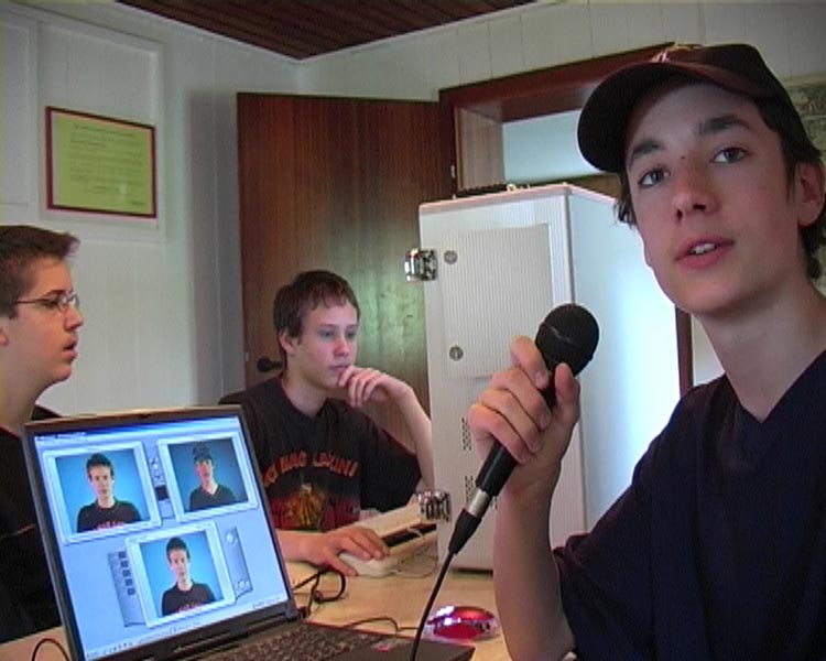 Projekt "Don´t believe what you see". Ein Junge sitzt vor einem Laptop und hält ein Mikrofon in der Hand. Auf dem Bildschirm des Laptops sind drei Portraits zu sehen. Im Hintergrund sitzen zwei weitere Jugendliche.