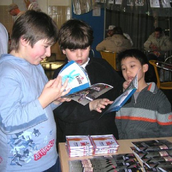 Projekt ""Eine Frage der Ehre?!" - Jugendmedienwettbewerb". Vor einem Tisch, auf dem Hefte und CDs ausgelegt sind, stehen drei Jungen. Zwei der Jungen haben jeweils eins der Hefte in der Hand und betrachten sie.