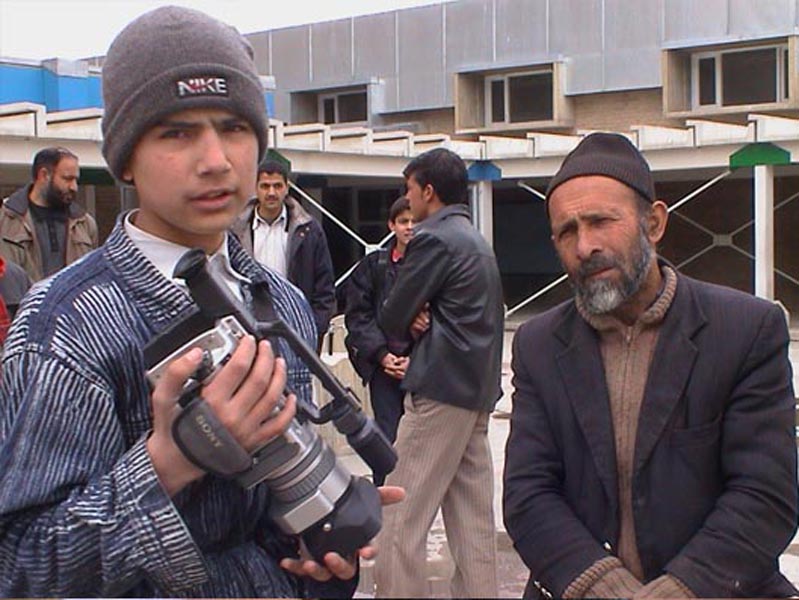 Projekt "Videobriefe Afghanistan Deutschland". Ein Junge, der eine Filmkamera in der Hand hält, steht neben einem älteren Herren. Im Hintergrund stehen weitere Personen in einer Gruppe zusammen.
