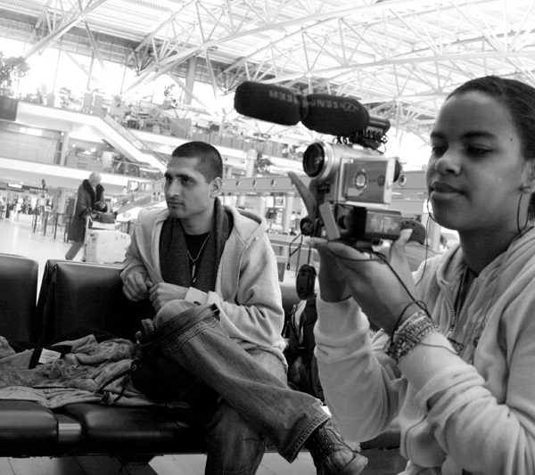 Projekt "ungeduldig". Schwarzweißfoto eines Flughafenwartebereichs. Ein Mädchen hält eine Kamera in der Hand, daneben sitzt ein junger Mann auf einer Bank.
