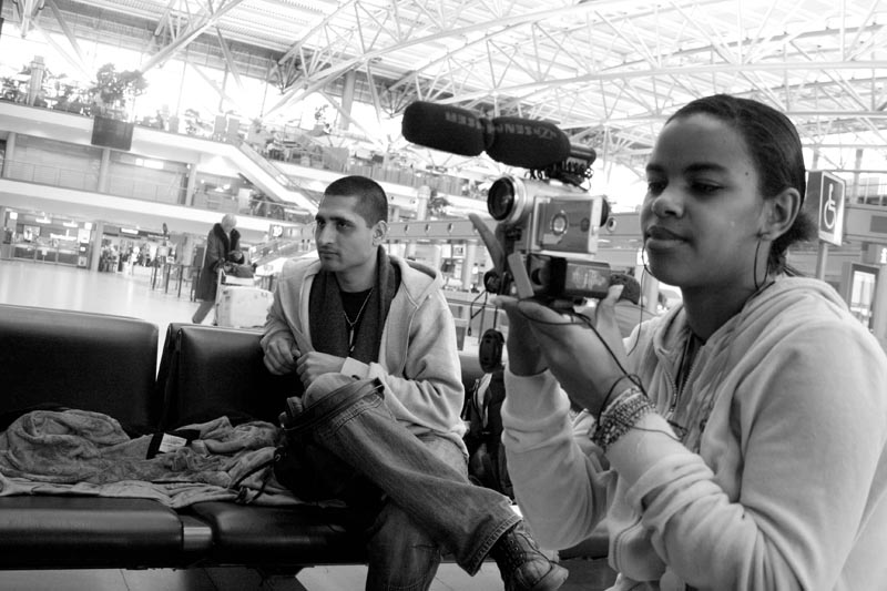 Projekt "ungeduldig". Schwarzweißfoto eines Flughafenwartebereichs. Ein Mädchen hält eine Kamera in der Hand, daneben sitzt ein junger Mann auf einer Bank.