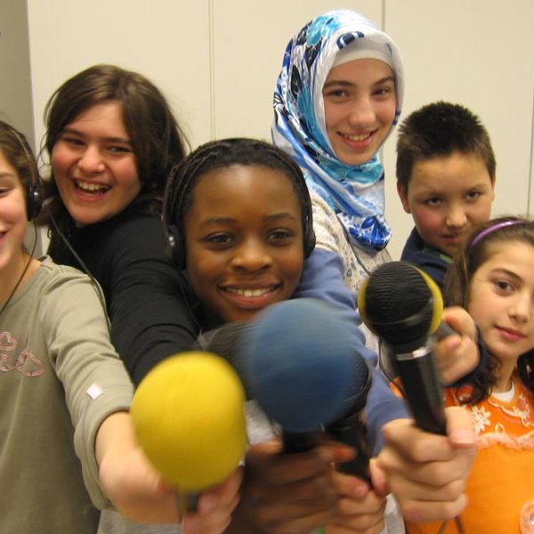 Projekt "Radiofüchse - Das interkulturelle Hamburger Kinderradio". Gruppenfoto einiger Mitwirkender des Projekts, die Mikrofone in die Kamera halten.