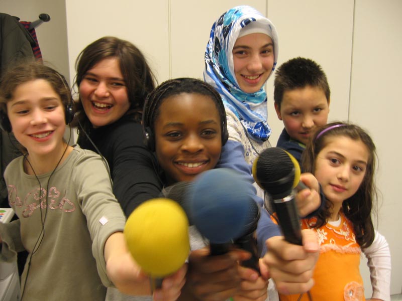 Projekt "Radiofüchse - Das interkulturelle Hamburger Kinderradio". Gruppenfoto einiger Mitwirkender des Projekts, die Mikrofone in die Kamera halten.