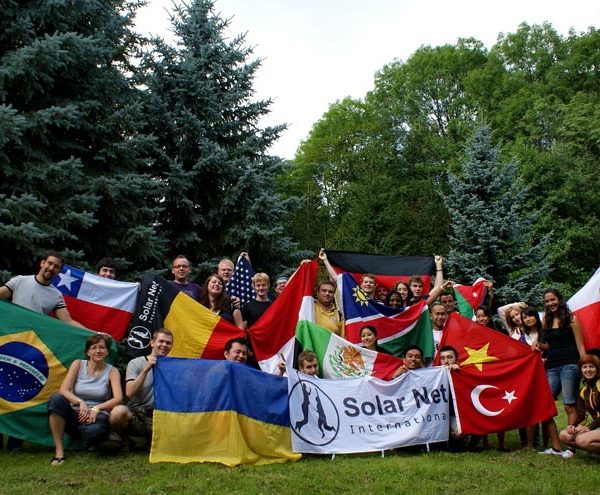 Projekt "Globales Lernen im Internet". Eine Gruppe von Personen posiert in einer parkähnlichen Kulisse mit verschiedenen Länderflaggen und einer Flagge mit der Aufschrift "Solar Net - International".