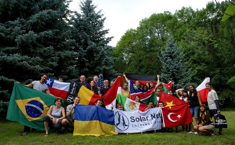 Projekt "Globales Lernen im Internet". Eine Gruppe von Personen posiert in einer parkähnlichen Kulisse mit verschiedenen Länderflaggen und einer Flagge mit der Aufschrift "Solar Net - International".