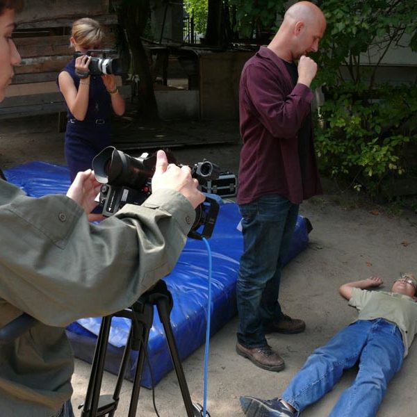 Projekt "5. Spinxx-Kritikergipfel FAKE IT!". Ein Junge liegt am Boden, neben ihm steht ein Erwachsener. Zwei weitere Personen halten Kameras, die auf die Szene gerichtet sind.