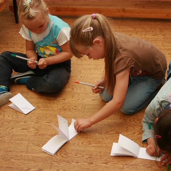 Projekt "Medienpädagogik im Vorschulalter". Drei Mädchen sitzen auf dem Boden und beschriften kleine Notizheftchen, die vor ihnen liegen.