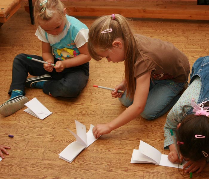 Projekt "Medienpädagogik im Vorschulalter". Drei Mädchen sitzen auf dem Boden und beschriften kleine Notizheftchen, die vor ihnen liegen.