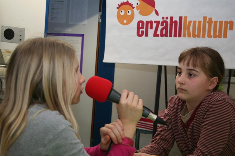 Projekt "erzählkultur - Sprachkompetenzförderung durch aktive Medienarbeit". Zwei junge Mädchen sitzen sich gegenüber. Eins von ihnen spricht in ein rotes Mikrofon.