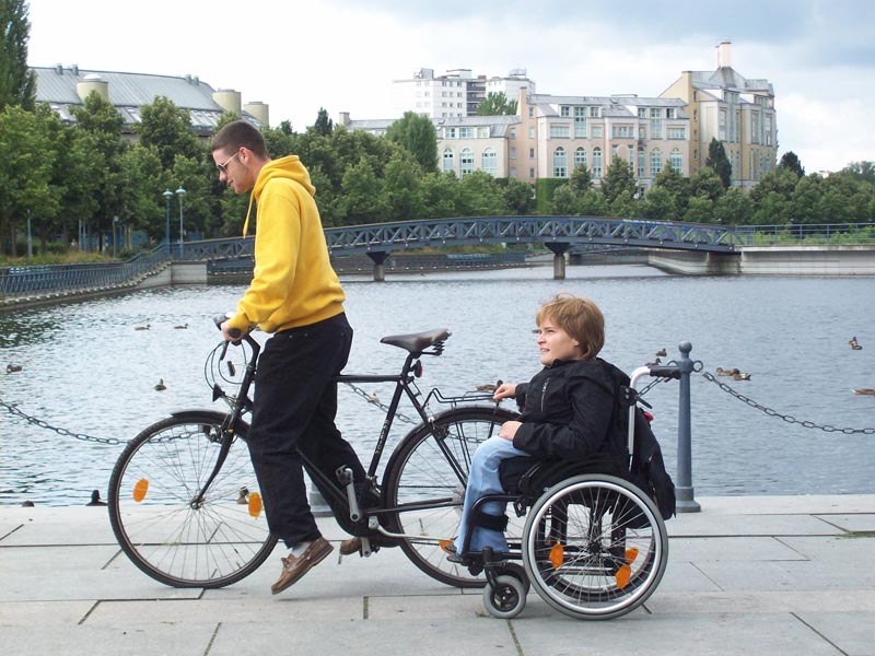 Projekt "Jugendfilmprojekt "Verfilmt in Berlin"". Ein junger Mann fährt ein Fahrrad, an dessen Gepäckträger sich eine Rollstuhlfahrerin festhält.