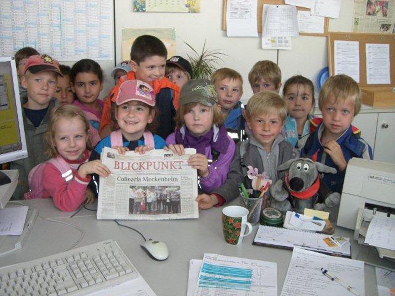 Projekt "Wie kommt unser Foto in die Zeitung?". Eine Gruppe Kinder steht hinter einem Schreibtisch und präsentiert eine Tageszeitung.