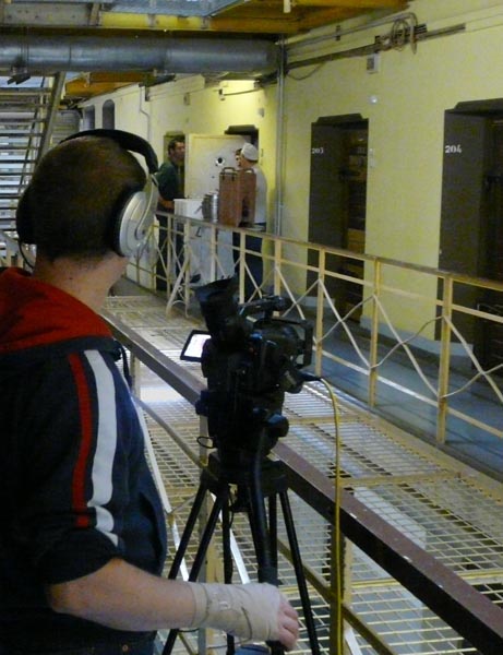 Projekt "podknast.de". In einem Gefängnisflur steht ein junger Mann hinter einer Filmkamera und trägt Kopfhörer.