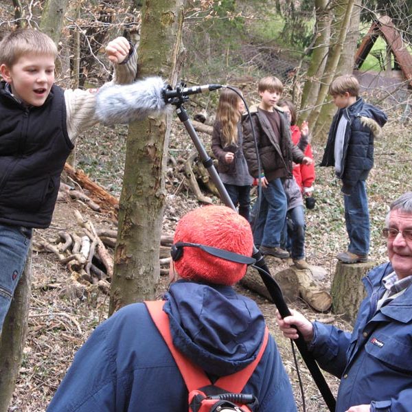 Projekt "Spiel- und Lieblingsorte: Ein Rundgang für die Ohren". In einem Wald stehen zwei mit Tonangel und Kopfhörern ausgestattete Erwachsene vor einem Jungen. Im Hintergrund steht eine Gruppe Kinder.