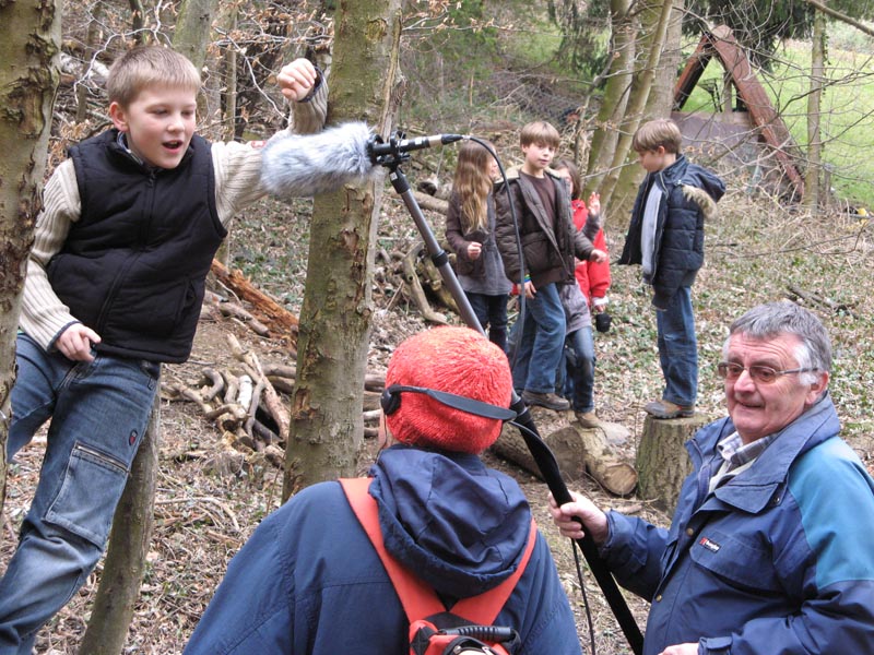 Projekt "Spiel- und Lieblingsorte: Ein Rundgang für die Ohren". In einem Wald stehen zwei mit Tonangel und Kopfhörern ausgestattete Erwachsene vor einem Jungen. Im Hintergrund steht eine Gruppe Kinder.