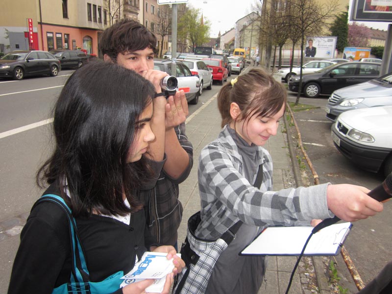 Projekt "LEONAU.TV - Das Web-TV für ST.LEOnhard & SchweiNau". Drei Jugendliche dsind mit Filmkamera, Mikrofon und Klemmbrett ausgestattet. Eine von ihnen streckt das Mikrofon einer Person entgegen, die außerhalb des Bildes steht.