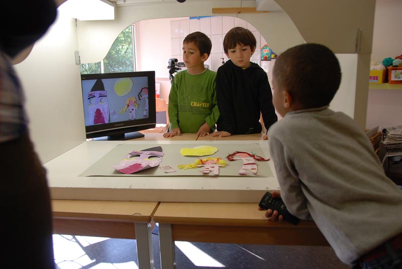 Projekt "Trickfilmprojekt "Die Abenteuer von Okarla und Jolise"". Um eine Trickbox herum stehen drei Kinder. Auf einem danebenstehenden Monitor ist das Bild aus der Trickbox abgebildet.