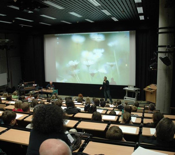 Projekt "Kinderfilmuniversität Potsdam Babelsberg". Aufnahme eines Hörsaals, in welchem überwiegend Kinder sitzen. Vorne vor der Leinwand steht eine Person und hält einen Vortrag.