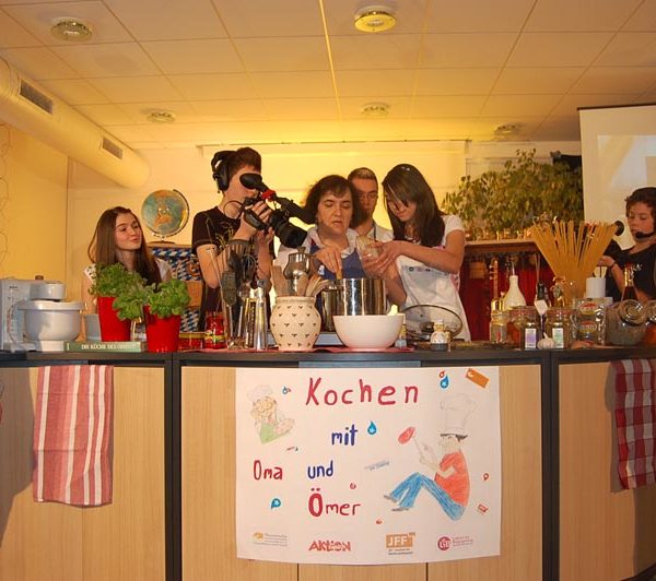 Projekt "Generationen im Dialog". Eine Gruppe Menschen steht um eine Küchenzeile. Eine ältere Dame steht hinter einem Kochtopf. Ein Jugendlicher filmt über ihre Schulter hinweg.