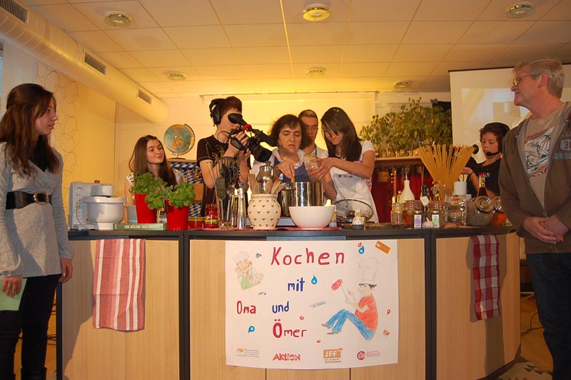 Projekt "Generationen im Dialog". Eine Gruppe Menschen steht um eine Küchenzeile. Eine ältere Dame steht hinter einem Kochtopf. Ein Jugendlicher filmt über ihre Schulter hinweg.
