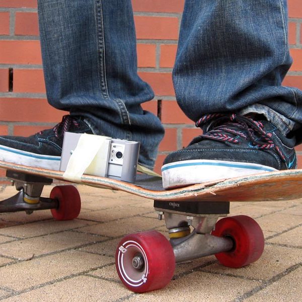 Projekt "MobileMovie - urbane Mobilität im künstlerischen Handyfilm". Großaufnahme eines Skateboards, auf dem eine Person steht. Zwischen den Füßen ist mit Klebeband ein Handy mit Kamera befestigt.