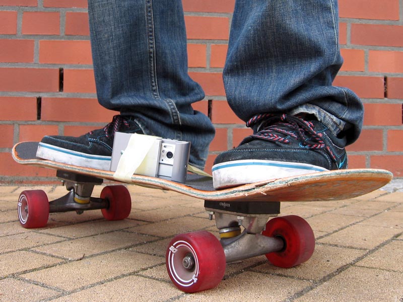 Projekt "MobileMovie - urbane Mobilität im künstlerischen Handyfilm". Großaufnahme eines Skateboards, auf dem eine Person steht. Zwischen den Füßen ist mit Klebeband ein Handy mit Kamera befestigt.