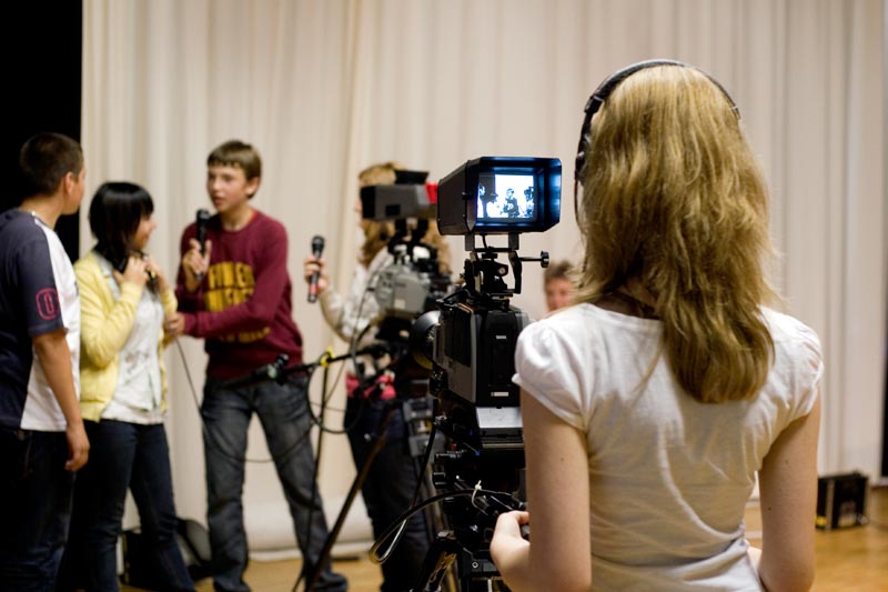 Projekt "Schulversuch "Schulische Medienbildung in Mecklenburg-Vorpommern"". Eine Jugendliche filmt mit einer Filmkamera vier weitere Jugendliche. Zwei von ihnen halten Mikrofone in der Hand.