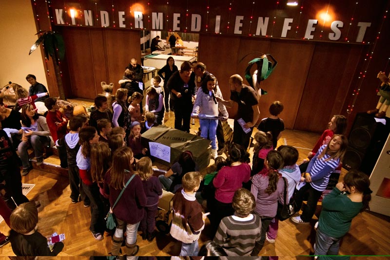 Projekt "Kindermedienfest - Expedition durch den Mediendschungel". Auf und um ein Podium steht eine Gruppe Kinder. In der Mitte auf dem Podium steht ein Erwachsener, der einem Kind neben ihm ein Mikrofon entgegenstreckt.