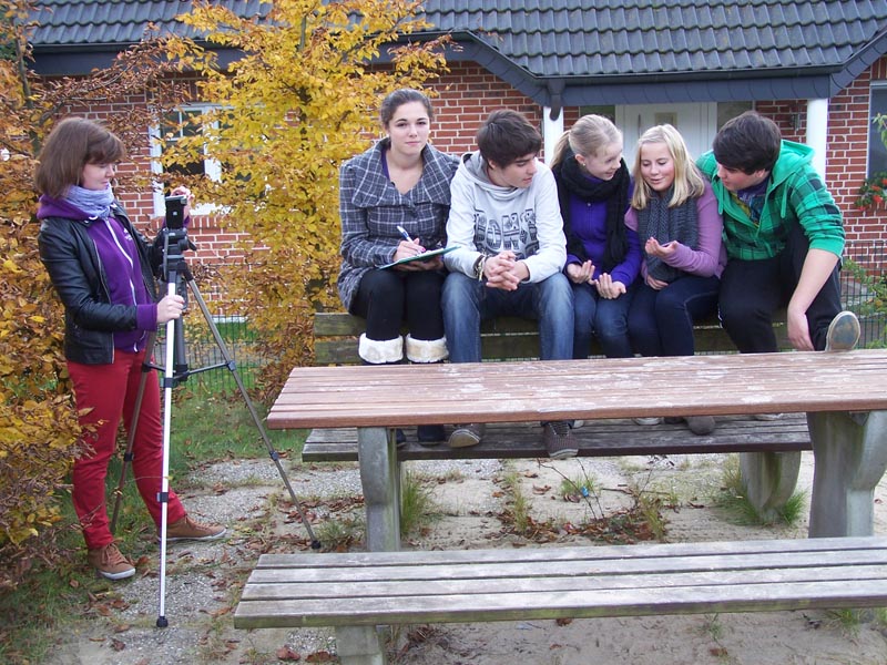 Projekt "Jäger des Vergessenen". Fünf Jugendliche sitzen auf der Lehne einer Bank, eine weitere steht mit einer Kamera daneben und filmt sie.