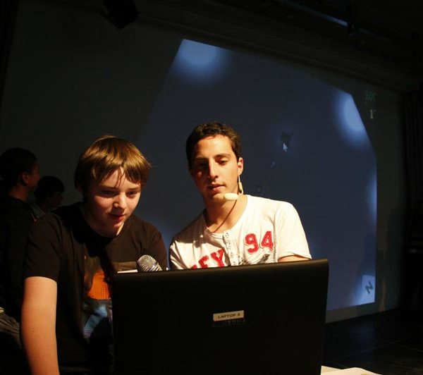 Projekt "MyyGames - Jugendtagungen zum Thema Computerspiele". Zwei junge Erwachsene, die mit Mikrofonen ausgestattet sind, stehen in einem dunklen Raum vor einem Laptop.