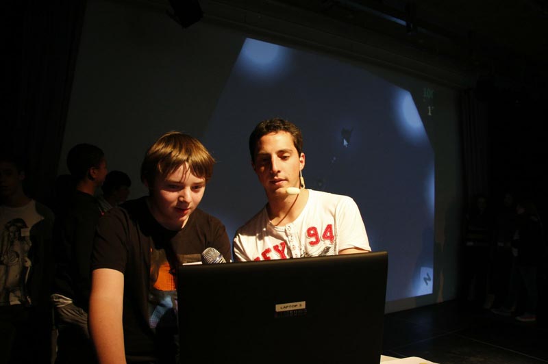 Projekt "MyyGames - Jugendtagungen zum Thema Computerspiele". Zwei junge Erwachsene, die mit Mikrofonen ausgestattet sind, stehen in einem dunklen Raum vor einem Laptop.