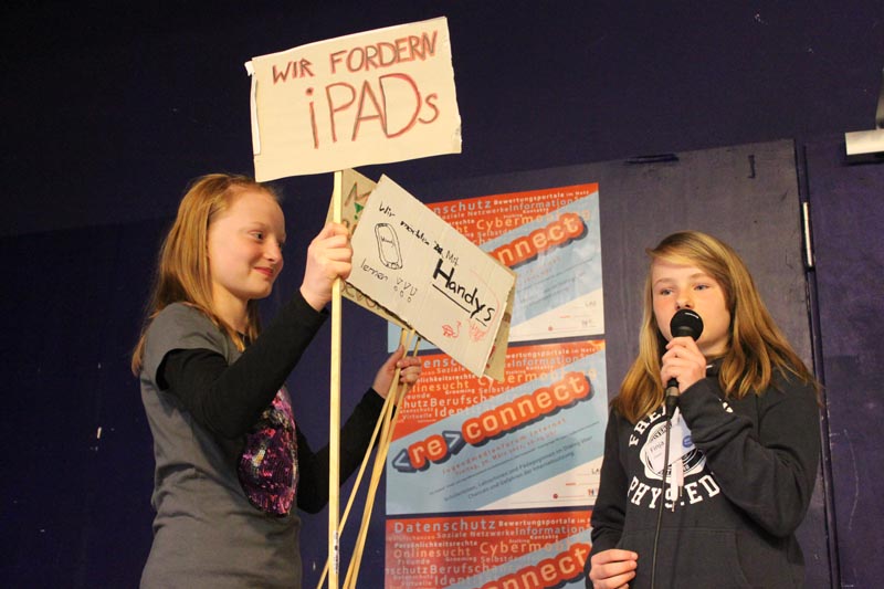 Projekt "connect Jugendmedienforum". Ein Mädchen hält Schilder in den Händen, auf denen "Wir fordern iPads" und "Wir möchten mit Handys lernen!!!" steht. Daneben steht ein weiteres und spricht in ein Mikrofon.
