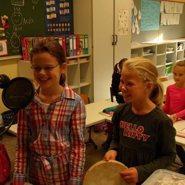 Projekt "Ohrenspitzer - Ein Projekt zur Zuhörförderung". Zwei Mädchen stehen in einem Klassenraum vor einem Mikrofon. Eins der Mädchen hält ein Tamburin in den Händen.