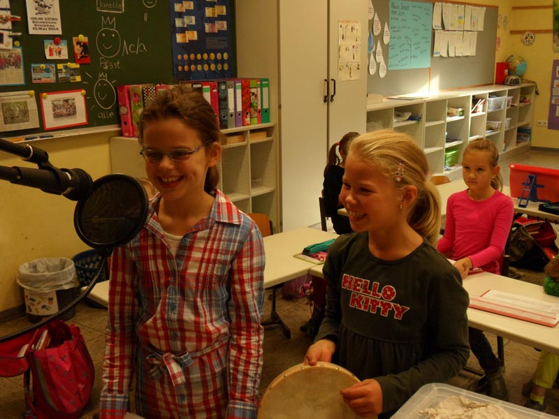 Projekt "Ohrenspitzer - Ein Projekt zur Zuhörförderung". Zwei Mädchen stehen in einem Klassenraum vor einem Mikrofon. Eins der Mädchen hält ein Tamburin in den Händen.