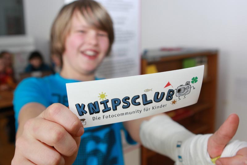 Projekt "Knipsclub - die Fotocommunity für Kinder von acht bis zwölf Jahren". Foto eines Jungen, der ein Schild mit em Logo des Knipsclubs in die Kamera hält.