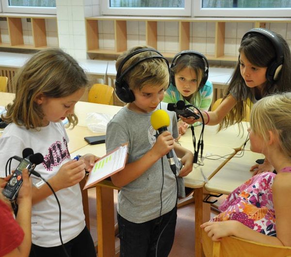 Projekt "Alle machen Radio! Live-Radio in der Grundschule". Foto einer Gruppe von Kindern. Einige von ihnen sind mit Kopfhörern und Mikrofonen ausgestattet.