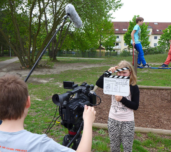 Projekt "(Cyber-)Mobbing - Aufgeklärt! Schüler der Stadt Cottbus klären auf". Auf einem Spielplatz stehen zwei Kinder auf einem der Spielgeräte. Drei weitere sind mit Tonangel, Kamera und Filmklappe ausgestattet und filmen die Szene.