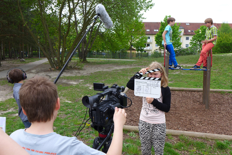 Projekt "(Cyber-)Mobbing - Aufgeklärt! Schüler der Stadt Cottbus klären auf". Auf einem Spielplatz stehen zwei Kinder auf einem der Spielgeräte. Drei weitere sind mit Tonangel, Kamera und Filmklappe ausgestattet und filmen die Szene.