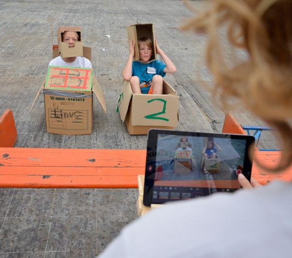 Projekt "TINKERTANK - Kreativ mit Technik". Foto von zwei Kindern, die in Pappkartons sitzen und mit einem Tablet fotografiert werden.