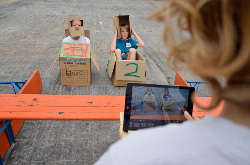 Projekt "TINKERTANK - Kreativ mit Technik". Foto von zwei Kindern, die in Pappkartons sitzen und mit einem Tablet fotografiert werden.