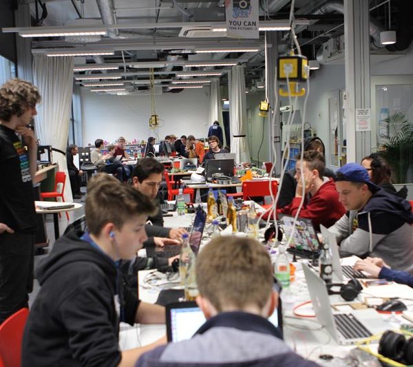 Projekt "Jugend hackt - Mit Code die Welt verbessern". Foto eines Raums mit vielen Tischen, an denen Jugendliche mit Laptops sitzen.