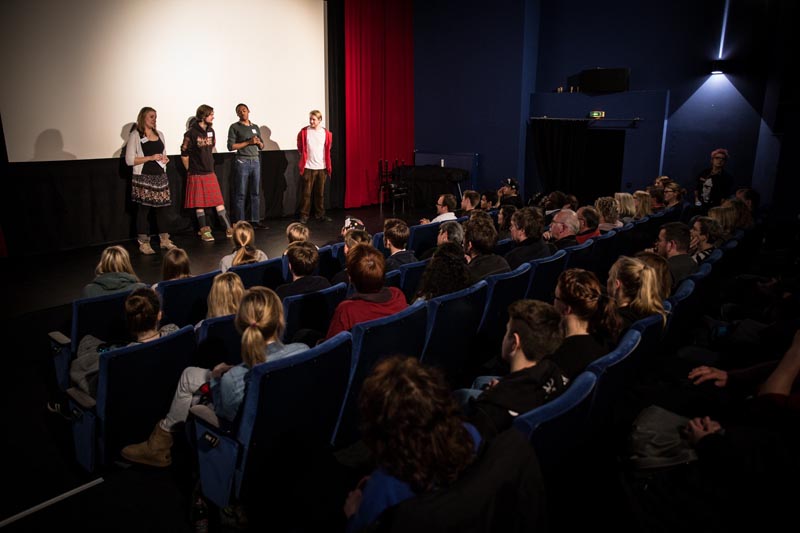 Projekt "Europa und du?! - Jugendfilmprojekt". Foto eines Kinosaals. Vorne vor der Leinwand stehen vier Personen.