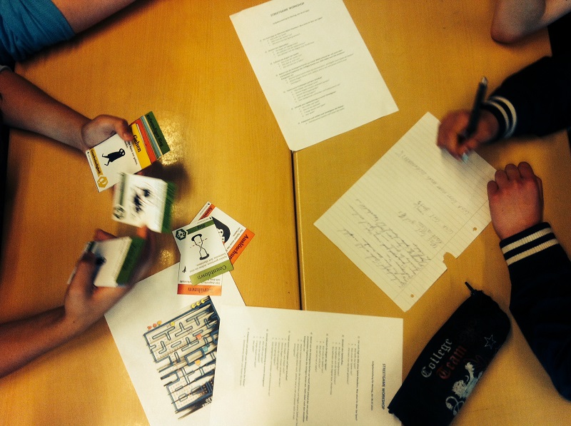 Projekt "Profilklasse Smart Gaming". Foto eines Tischs, auf dem Zettel liegen. Eine der Personen, die am Tisch sitzen hält Spielkarten in der Hand, eine andere schreibt auf einen Zettel.