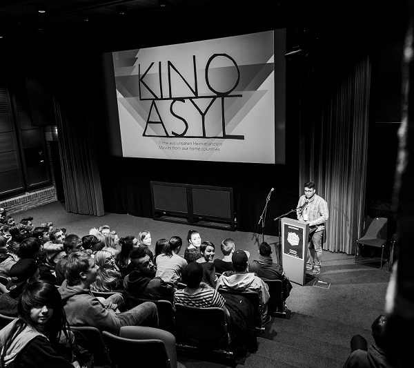Projekt "Kino Asyl". Foto eines Saals. Im Vordergrund ein Teil des Publikums, im Hintergrund eine Person, die in ein Mikrofon spricht und eine Leinwand mit dem Logo des Projekts.