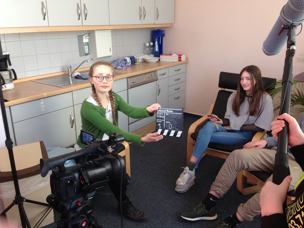 Projekt "Durchblick im Netz". In einer Küche sitzen zwei Jugendliche vor einer Kamera. Eine weitere hält eine Filmklappe vor die Kamera.