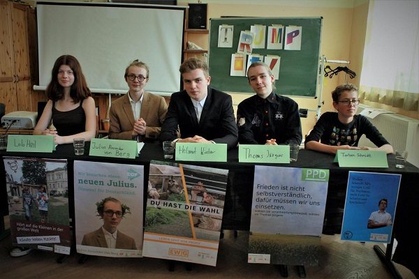 Projekt "TRUMP IT! Medienwahlkampf macht Schule". An einem Tisch sitzen fünf Jugendliche nebeneinander. Vor ihnen stehen Namensschilder und am Tisch hängen Wahlplakate.